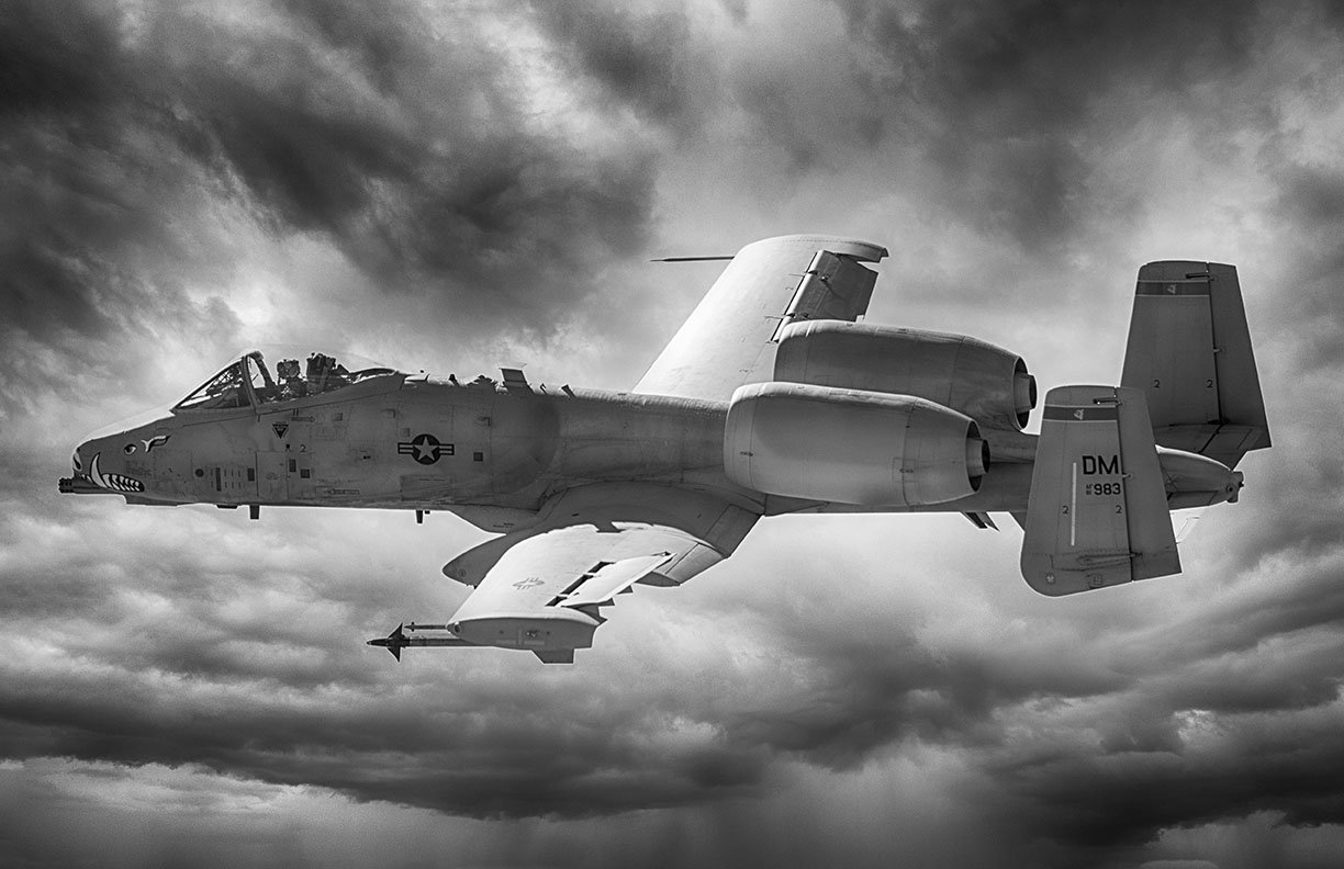 Black and white dramatic composite photograph of an A-10 Warthog Thunderbolt II in flight, viewed from below against powerful storm clouds. Iconic shark mouth nose art, USAF roundel, and tail markings