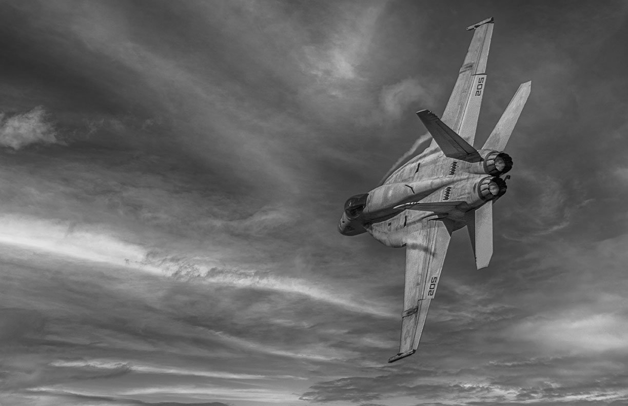 Black-and-white composite photograph of an F/A-18 Hornet banking through dramatic clouds with visible vapor trails, showcasing modern military aviation in fine art.