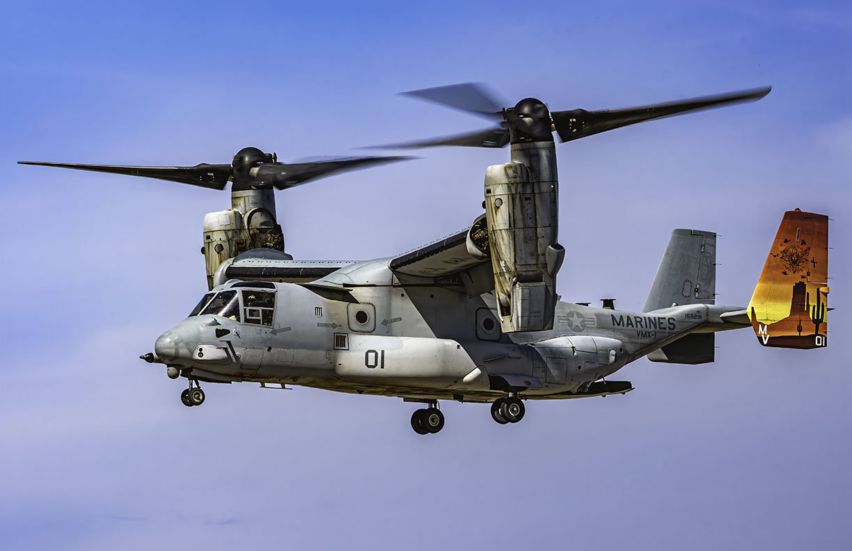 A high-resolution photograph of a United States Marine Corps MV-22B Osprey tiltrotor aircraft from the VMX-1 squadron in flight, showcasing its large propellers and military markings against the sky. 